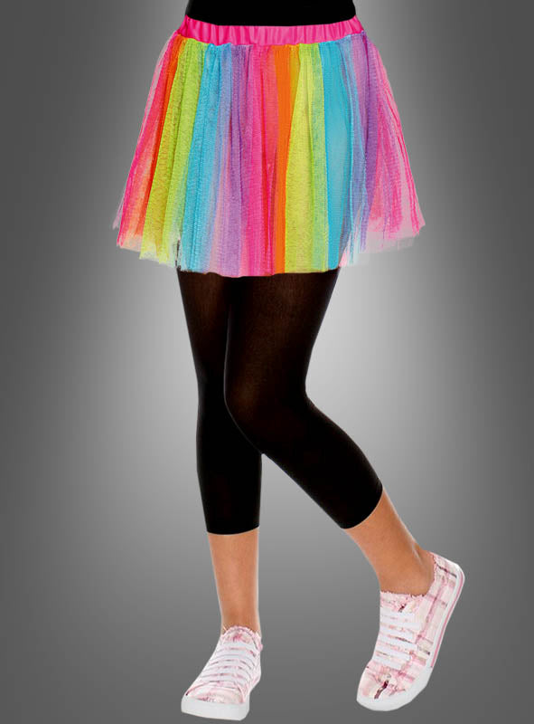 Eine Person, die einen „Bunter Tüllrock Regenbogen Kinder“ trägt, dazu schwarze Leggings und rosa Turnschuhe, steht vor einem neutralen grauen Hintergrund.