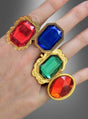 Eine Hand hält vier „Goldene Ringe mit Edelstein, verstellbar“ in die Hand, die jeweils mit großen roten, blauen, grünen und orangefarbenen Edelsteinen besetzt sind – darunter ein auffälliger Edelsteinring – vor einem grau verlaufenden Hintergrund. Ideal als Kostümschmuck für den Karneval oder um jedem Anlass das gewisse Etwas zu verleihen.