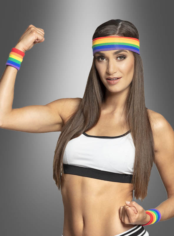 Eine Frau, die einen Sport-BH, ein Regenbogen-Stirnband und das Schweißband-Set „Regenbogen CSD“ trägt, spannt ihren Arm an und lächelt vor einem grauen Hintergrund mit Farbverlauf – dabei strahlt sie eine Energie aus, die perfekt zum Christopher Street Day oder zu jeder Pride-Feier passt.