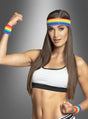 Eine Frau, die einen Sport-BH, ein Regenbogen-Stirnband und das Schweißband-Set „Regenbogen CSD“ trägt, spannt ihren Arm an und lächelt vor einem grauen Hintergrund mit Farbverlauf – dabei strahlt sie eine Energie aus, die perfekt zum Christopher Street Day oder zu jeder Pride-Feier passt.