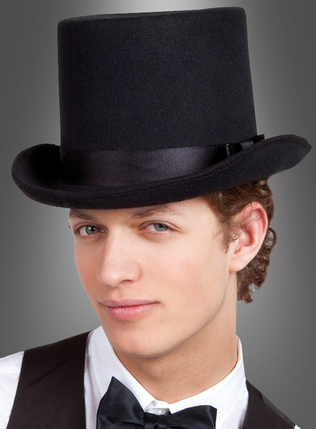 Ein junger Mann mit einem „Schwarzer Zylinder Hut Deluxe“ und einer Fliege, gekleidet in eine stilvolle schwarze Weste, die sein Zauberer-Kostüm perfekt ergänzt, blickt mit einem leichten Lächeln vor einem grauen Hintergrund in die Kamera.