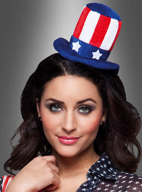 Eine Frau mit dunklem Haar trägt einen kleinen rot-weiß-blauen USA-Minizylinder mit Haarreif im Uncle-Sam-Stil und ein Shirt mit Sternenmuster, wodurch vor grauem Hintergrund ein verspieltes Uncle-Sam-Outfit entsteht.