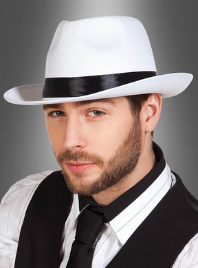 Ein Mann mit kurzem Bart trägt den „Weißen Gangsterhut Mafia“ – einen weißen Fedora mit schwarzem Band –, dazu ein gestreiftes Hemd, eine schwarze Weste und eine schwarze Krawatte, und posiert vor einem grau verlaufenden Hintergrund, um den authentischen Look eines Mafiakostüms aus den 20er Jahren zu verkörpern.