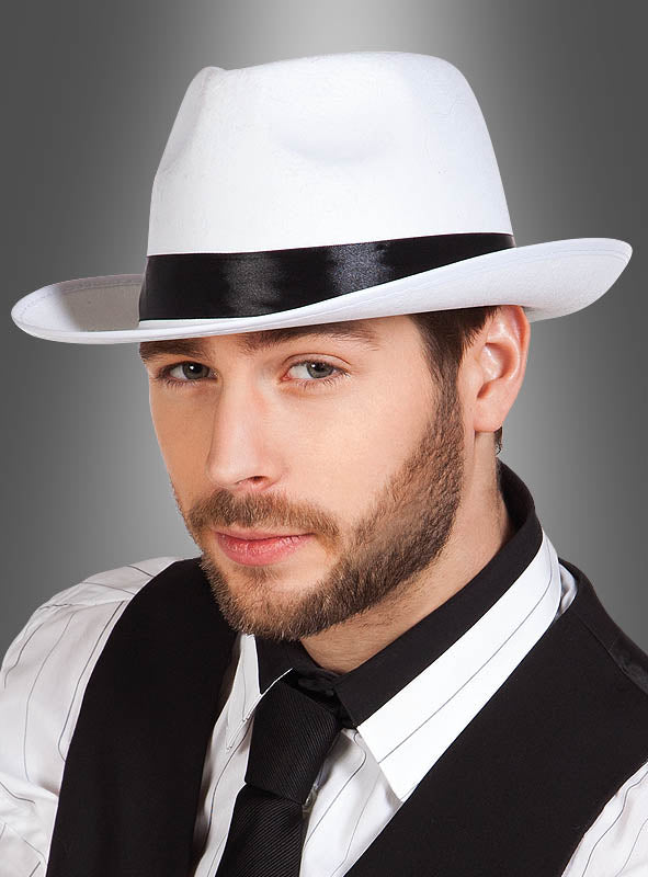 Ein Mann mit kurzem Bart trägt den „Weißen Gangsterhut Mafia“ – einen weißen Fedora mit schwarzem Band –, dazu ein gestreiftes Hemd, eine schwarze Weste und eine schwarze Krawatte, und posiert vor einem grau verlaufenden Hintergrund, um den authentischen Look eines Mafiakostüms aus den 20er Jahren zu verkörpern.