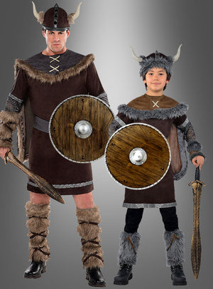 Ein Erwachsener und ein Kind, als Wikinger verkleidet, stehen vor einem grauen Hintergrund mit Farbverlauf. Beide halten jeweils ein „Rundschild 40 cm in Holzoptik“ – authentische runde Schilde im Holzlook, die von traditionellen Rundschild-Designs inspiriert sind – sowie Schwerter in der Hand; beide tragen Hörnerhelme und mit Kunstfell besetzte Kostüme.
