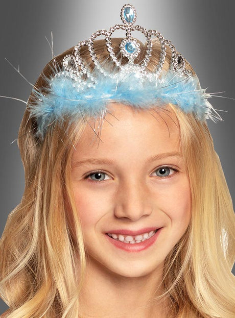 Ein junges Mädchen mit langen blonden Haaren lächelt vor einem grauen Hintergrund mit Farbverlauf und trägt dabei die „Eisprinzessin“-Krone für Kinder – eine funkelnde silberne Krone, die mit blauen Edelsteinen und flauschigem blauem Besatz verziert ist.
