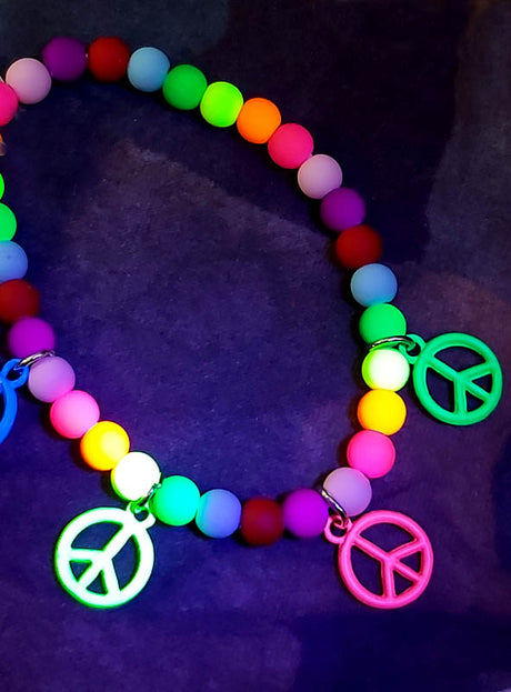 Das „Bunte Hippiearmband mit Peace-Zeichen“ ist ein einzigartiges Hippie-Schmuckstück, das mit bunten Perlen und drei Anhängern in Form von Peace-Zeichen verziert ist und eindrucksvoll vor einem dunklen Hintergrund fotografiert wurde.