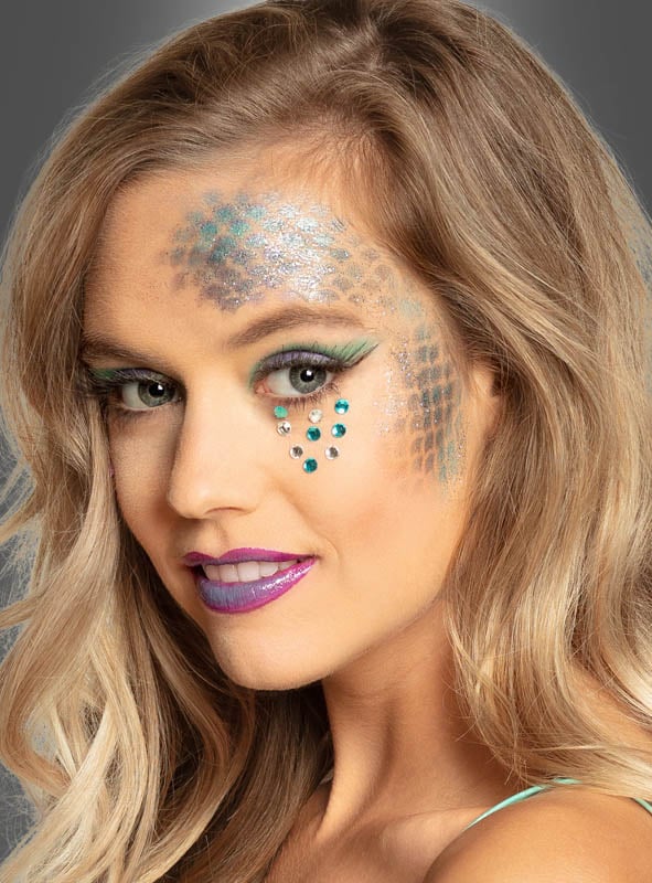 Eine Frau mit welligem blondem Haar trägt auf der Stirn ein schillerndes, von Fischschuppen inspiriertes Make-up, das mit dem Glitzer-Gel aus dem Schminkset „Meerjungfrau“ aufgetragen wurde. Akzente setzen grüne Strasssteine unter dem Auge, ein kräftiger violetter Lippenstift und schimmernder grüner Lidschatten.