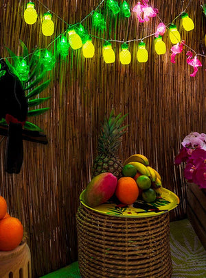 Auf einem Korb-Tisch sind verschiedene tropische Früchte unter der Lichterkette „Ananas“ 140 cm „Sommerparty“ ausgestellt, während rosa Blumendekorationen die festliche „Sommerparty“-Dekoration vor einer Bambuswand unterstreichen – perfekt für jede Hawaii-Party.