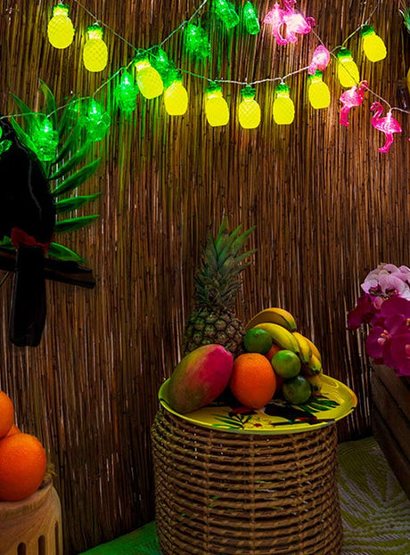 Auf einem Korb-Tisch sind verschiedene tropische Früchte unter der Lichterkette „Ananas“ 140 cm „Sommerparty“ ausgestellt, während rosa Blumendekorationen die festliche „Sommerparty“-Dekoration vor einer Bambuswand unterstreichen – perfekt für jede Hawaii-Party.