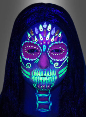 Eine Person mit langen, dunklen Haaren und einem Gesicht, das mit dem UV-Schmink-Set für Kinder und Erwachsene mit fluoreszierenden, tötenähnlichen Motiven bemalt ist und unter Schwarzlicht vor einem grauen Hintergrund lebhaft leuchtet.