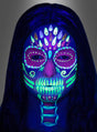 Eine Person mit langen, dunklen Haaren und einem Gesicht, das mit dem UV-Schmink-Set für Kinder und Erwachsene mit fluoreszierenden, tötenähnlichen Motiven bemalt ist und unter Schwarzlicht vor einem grauen Hintergrund lebhaft leuchtet.