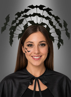 Eine Frau lächelt vor einem grauen Hintergrund. Sie trägt einen schwarzen Umhang, Fledermaus-Gesichtssticker und den Haarreif „Fledermaus-Schwarm“ – ein perfektes Halloween-Kostüm-Accessoire für jede gruselige Feier.