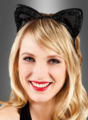 Eine Frau mit blonden Haaren und rotem Lippenstift lächelt vor einem grauen Hintergrund mit Farbverlauf in die Kamera. Sie trägt einen schwarzen, glitzernden Haarreif mit Katzenohren – das perfekte Accessoire für jeden verspielten Katzenkostüm-Look.
