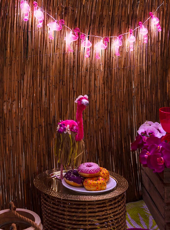 Auf einem kleinen runden Korb-Tisch stehen ein Teller mit bunten Donuts und eine rosa Flamingo-Dekoration, während im Hintergrund die „Lichterkette 140 cm Flamingo Sommerparty“ mit leuchtenden LED-Lichtern hängt – perfekt für deine nächste Sommerparty.