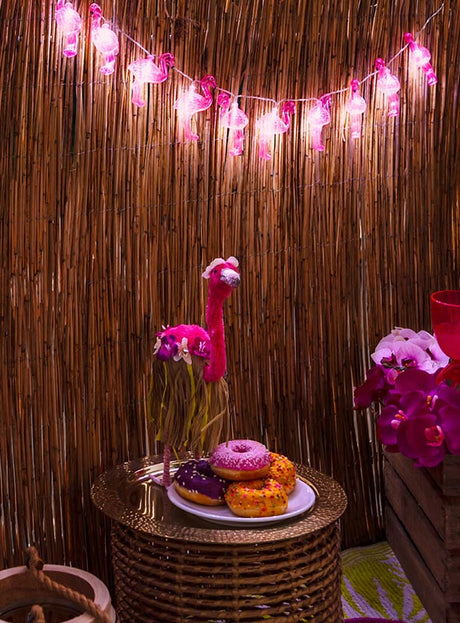 Auf einem kleinen runden Korb-Tisch stehen ein Teller mit bunten Donuts und eine rosa Flamingo-Dekoration, während im Hintergrund die „Lichterkette 140 cm Flamingo Sommerparty“ mit leuchtenden LED-Lichtern hängt – perfekt für deine nächste Sommerparty.