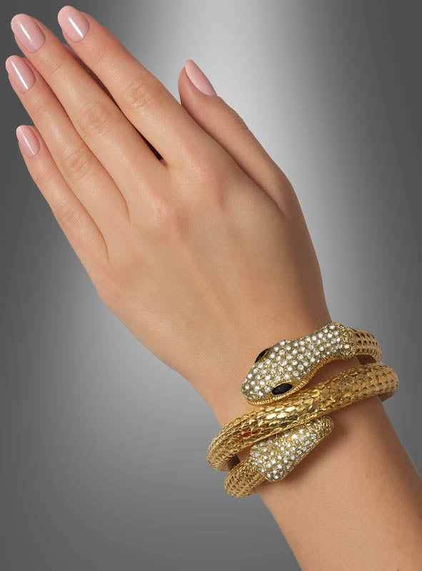 Eine Hand mit manikürten Nägeln trägt einen goldenen Schlangen-Armschmuck – ein exquisites, schlangenförmiges Armband, das mit klaren Steinen besetzt ist und zwei Schlangenköpfe mit dunklen Augen aufweist –, das an die Eleganz eines Kleopatra-Accessoires erinnert und vor einem grau abgestuften Hintergrund zur Geltung kommt.