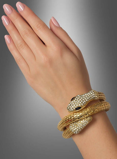 Eine Hand mit manikürten Nägeln trägt einen goldenen Schlangen-Armschmuck – ein exquisites, schlangenförmiges Armband, das mit klaren Steinen besetzt ist und zwei Schlangenköpfe mit dunklen Augen aufweist –, das an die Eleganz eines Kleopatra-Accessoires erinnert und vor einem grau abgestuften Hintergrund zur Geltung kommt.