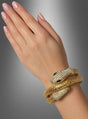 Eine Hand mit manikürten Nägeln trägt einen goldenen Schlangen-Armschmuck – ein exquisites, schlangenförmiges Armband, das mit klaren Steinen besetzt ist und zwei Schlangenköpfe mit dunklen Augen aufweist –, das an die Eleganz eines Kleopatra-Accessoires erinnert und vor einem grau abgestuften Hintergrund zur Geltung kommt.
