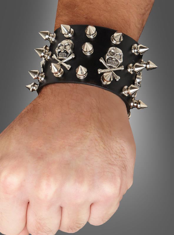Eine Hand, an der das „Punk Armband schwarz deluxe“ – ein schwarzes Lederarmband, verziert mit silbernen Nieten und Totenköpfen – trägt, hebt sich deutlich von einem grauen Hintergrund ab.
