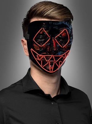 Eine Person, die ein schwarzes Hemd und die „LED Killer Maske Purger“ trägt – eine schwarze Maske mit roten LED-Linien, die Augen, Nase und den gezackten Mund umrahmen –, steht vor einem grauen Hintergrund.
