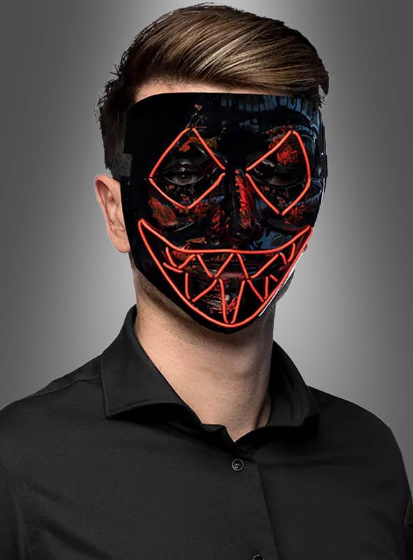 Eine Person, die ein schwarzes Hemd und die „LED Killer Maske Purger“ trägt – eine schwarze Maske mit roten LED-Linien, die Augen, Nase und den gezackten Mund umrahmen –, steht vor einem grauen Hintergrund.