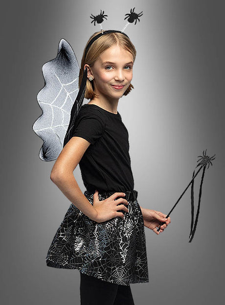 Ein junges Mädchen posiert selbstbewusst vor einem grauen Hintergrund und trägt das „Spinnen-Kostüm-Set für Kinder“ – ein Outfit mit schwarzen Spinnennetzflügeln, einem Stirnband mit Spinnen und einem passenden Zauberstab. Das perfekte Halloween-Kostüm für Mädchen!.