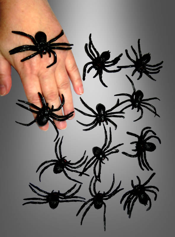 Eine Hand, auf der mehrere „Schwarze Spinnen aus Gummi“ (6 cm) aus der 12er-Packung liegen, umgeben von weiteren identischen Spinnenspielzeugen auf grauem Hintergrund – perfekt als Halloween-Dekoration.