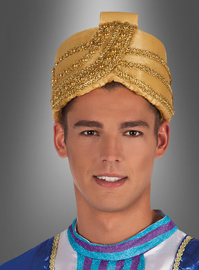 Ein Mann, der einen goldenen, mit Edelsteinen verzierten „Goldener Wüstenprinz“-Turban und ein blaues, goldbesetztes „Turban-Sultan“-Kostüm trägt, steht vor einem grauen Hintergrund mit Farbverlauf.