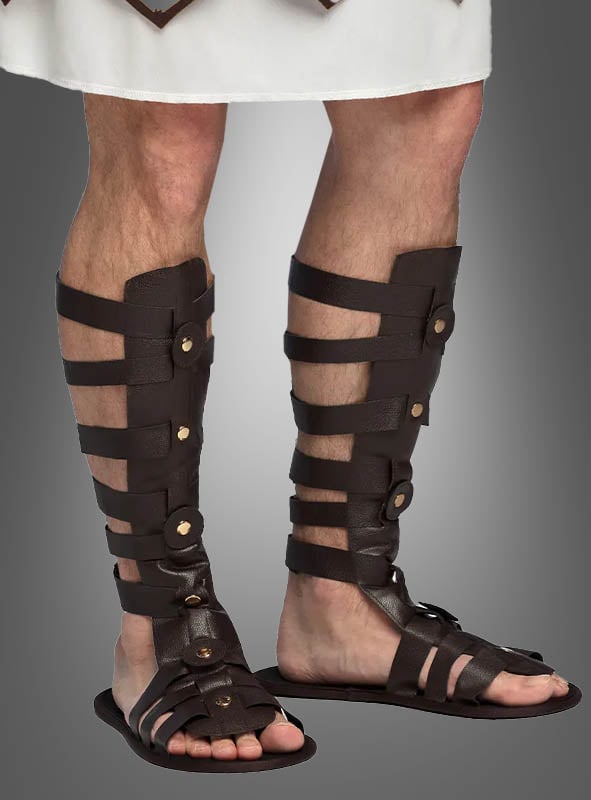 Eine Person, die dunkelbraune Gladiator-Sandalen im Römer-Stil und ein weißes Gewand trägt und vor einem grauen Hintergrund steht, verkörpert mühelos die Essenz eines antiken Kostüm-Accessoires.