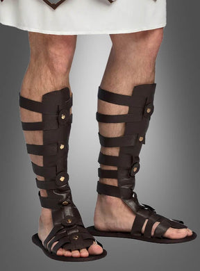 Eine Person, die dunkelbraune Gladiator-Sandalen im Römer-Stil und ein weißes Gewand trägt und vor einem grauen Hintergrund steht, verkörpert mühelos die Essenz eines antiken Kostüm-Accessoires.