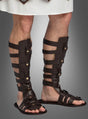 Eine Person, die dunkelbraune Gladiator-Sandalen im Römer-Stil und ein weißes Gewand trägt und vor einem grauen Hintergrund steht, verkörpert mühelos die Essenz eines antiken Kostüm-Accessoires.