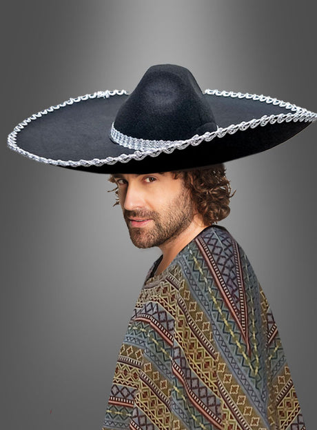 Ein Mann mit lockigem Haar trägt einen großen schwarzen Sombrero und einen gemusterten Poncho und steht vor einem einfarbigen grauen Hintergrund mit Farbverlauf.