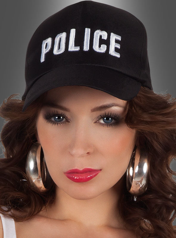 Eine Frau mit langen braunen Haaren, die eine „Polizei“-Mütze – eine schwarze Mütze mit der Aufschrift „POLICE“ – sowie große Creolen und Make-up trägt, blickt vor einem grauen Hintergrund direkt in die Kamera.