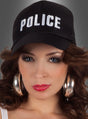 Eine Frau mit langen braunen Haaren, die eine „Polizei“-Mütze – eine schwarze Mütze mit der Aufschrift „POLICE“ – sowie große Creolen und Make-up trägt, blickt vor einem grauen Hintergrund direkt in die Kamera.