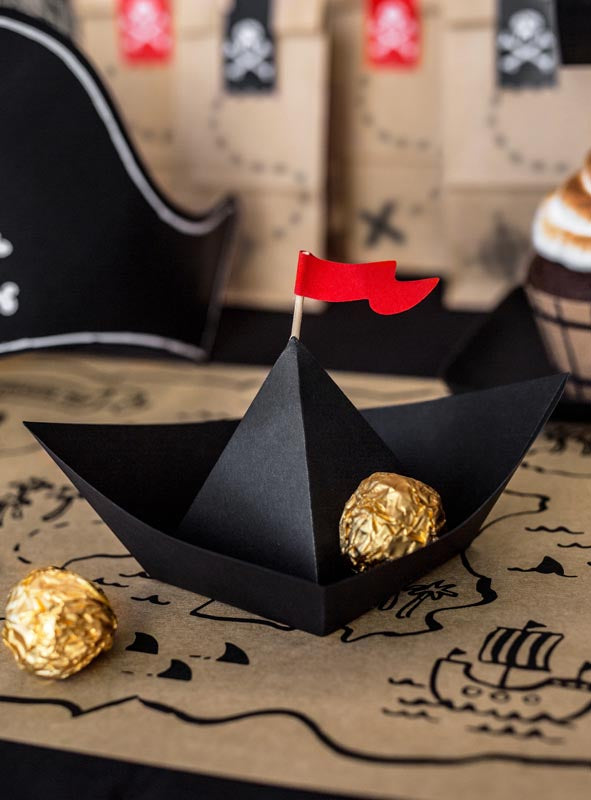 Eine schwarze Piratenschiff-Dekoration aus Pappe aus dem 6er-Pack, komplett mit roter Flagge und goldverpackten Pralinen, thront auf einer Karte im Piraten-Design und ist umgeben von Piraten-Partydekoration wie einem Piratenhut und verzierten Tüten – die perfekte Inspiration für deinen nächsten Piraten-Kindergeburtstag.