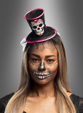 Eine Person, die einen kleinen „Tag der Toten“-Mini-Zylinder trägt und deren Gesicht mit aufwendigen Totenkopf- und Blumenmotiven im Stil des „Tag der Toten“ bemalt ist, steht vor einem neutralen Hintergrund.