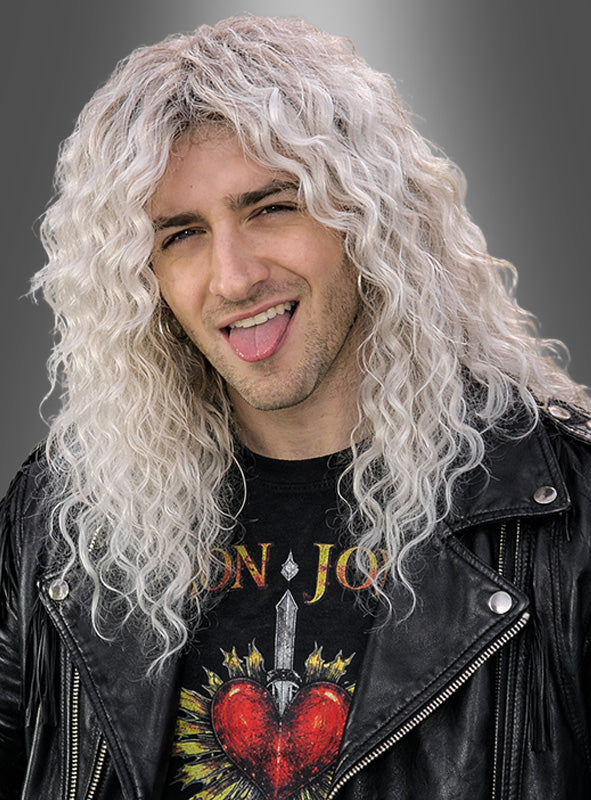 Eine Person mit langen, lockigen, platinblonden Haaren, die zu einer „Rocker-Perücke“ mit weißen Strähnen frisiert sind, trägt eine schwarze Lederjacke und ein Bon-Jovi-T-Shirt, streckt die Zunge heraus und posiert vor einem grauen Hintergrund.