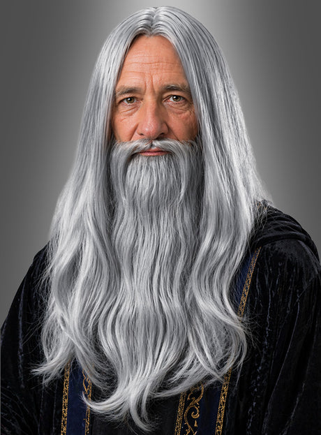 Ein älterer Mann mit langem grauen Bart und grauen Haaren trägt eine dunkle Robe mit goldenen Verzierungen und posiert vor einem grauen Hintergrund – die perfekte Inspiration für alle, die die „Perücke und Bart Merlin Zauberer“ in Betracht ziehen oder den faszinierenden Look dieser beeindruckenden grauen Langhaarperücke besonders hervorheben möchten.