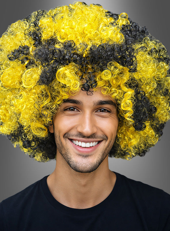 Vor einem neutralen grauen Hintergrund trägt eine lächelnde Person die Afro-Perücke „Biene“ in Schwarz-Gelb, eine große, lockige Perücke, die an eine Hummel erinnert.