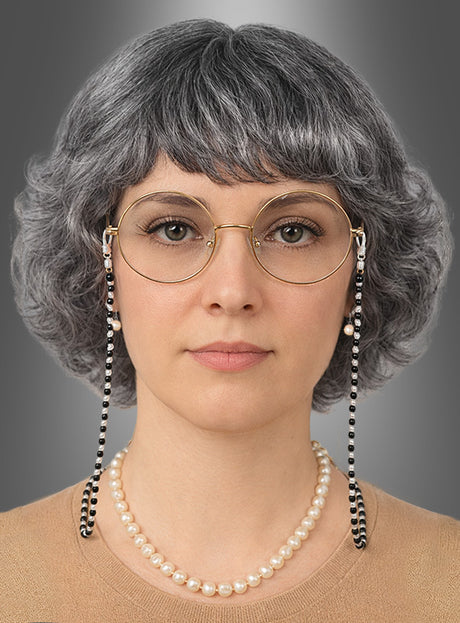 Eine Frau mit kurzem, grauem Haar, das als Perücke aus dem 4-teiligen „Oma-Perücken-Set“ gestylt ist, trägt eine runde Brille an einer Kette, ein beiges Oberteil und eine Perlenkette aus einem „Oma-Schmuck-Set“ und blickt vor einem grauen Hintergrund mit Farbverlauf in die Kamera.
