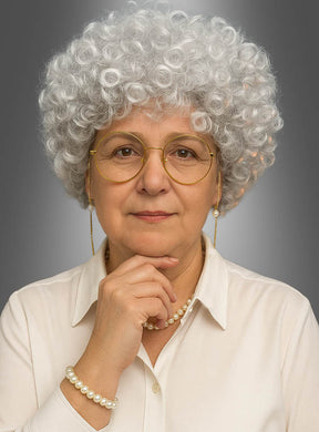 Eine ältere Frau mit kurzen, lockigen grauen Haaren, die mit der „Silbergrauen Oma-Perücke Golden Girl mit Zubehör“ frisiert sind, trägt eine Brille, eine Perlenkette und ein Perlenarmband sowie eine cremefarbene Bluse. Vor einem neutralen grauen Hintergrund blickt sie mit der Hand am Kinn in die Kamera.