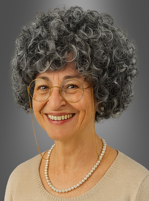 Eine lächelnde ältere Frau mit grauen Locken, einer runden Brille mit Kette, einer Perlenkette und einem beigen Oberteil vor grauem Hintergrund – ideal, um das „Oma graue Lockenperücke mit Schmuck“-Set als Teil eines authentischen Omakostüms zu präsentieren.