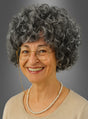 Eine lächelnde ältere Frau mit grauen Locken, einer runden Brille mit Kette, einer Perlenkette und einem beigen Oberteil vor grauem Hintergrund – ideal, um das „Oma graue Lockenperücke mit Schmuck“-Set als Teil eines authentischen Omakostüms zu präsentieren.