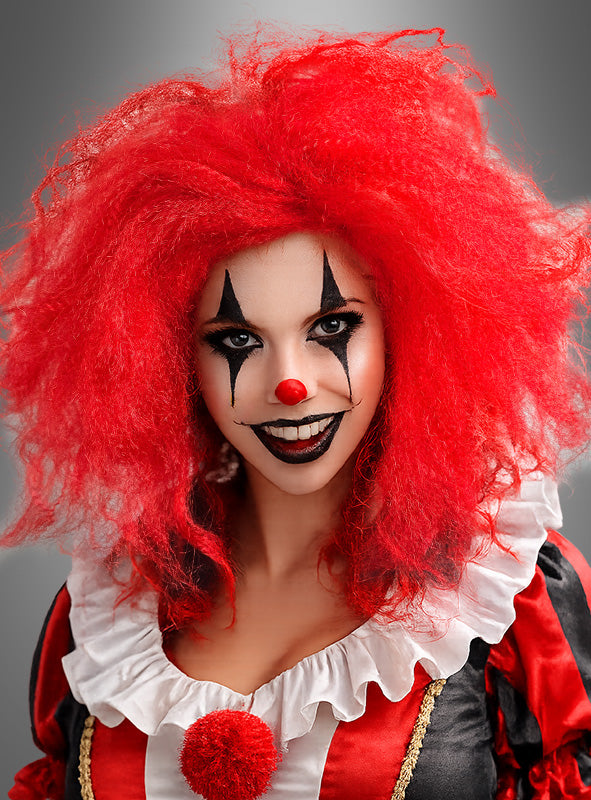Eine als Clown verkleidete Person mit einer leuchtend roten, gekräuselten Lockenperücke, weißer Gesichtsbemalung, schwarzem Augen- und Lippen-Make-up, einer roten Nase, einem Rüschenkragen und einem schwarz-roten Kostüm vor einem grauen Hintergrund.