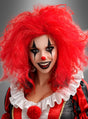Eine als Clown verkleidete Person mit einer leuchtend roten, gekräuselten Lockenperücke, weißer Gesichtsbemalung, schwarzem Augen- und Lippen-Make-up, einer roten Nase, einem Rüschenkragen und einem schwarz-roten Kostüm vor einem grauen Hintergrund.