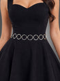 Eine Frau trägt ein ärmelloses, schwarzes Kleid mit Herzausschnitt, das mit einem silbernen Retro-Hüftgürtel kombiniert ist und an den Look eines Outfits aus den 60er- und 70er-Jahren erinnert. Vor einem einfarbigen grauen Hintergrund ist nur ihr Oberkörper zu sehen.