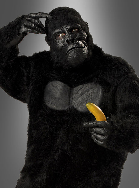 Eine Person, die das Gorilla-Kostüm für Herren in Größe XL trägt, kratzt sich am Kopf und zeigt auf eine Banane, während sie vor einem schlichten Hintergrund mit Farbverlauf steht.