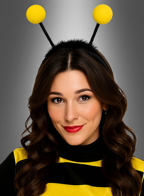 Eine Frau mit langen braunen Haaren trägt ein Bienenkostüm, bestehend aus einem gelb-schwarz gestreiften Oberteil und dem Stirnband „Haarreif Biene mit LED-Fühlern“ mit funkelnden schwarzen LED-Fühlern, an deren Enden gelbe Kugeln angebracht sind, vor einem grauen Hintergrund mit Farbverlauf.