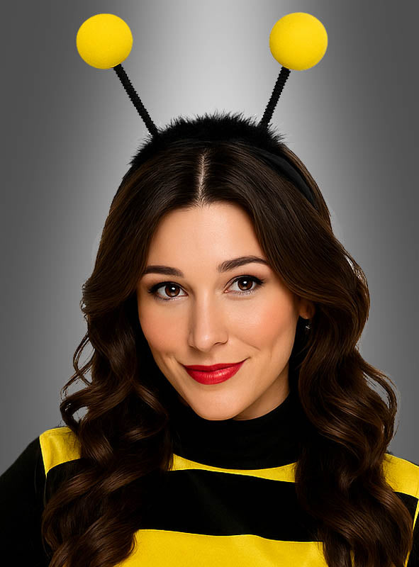 Eine Frau mit langen braunen Haaren trägt ein Bienenkostüm, bestehend aus einem gelb-schwarz gestreiften Oberteil und dem Stirnband „Haarreif Biene mit LED-Fühlern“ mit funkelnden schwarzen LED-Fühlern, an deren Enden gelbe Kugeln angebracht sind, vor einem grauen Hintergrund mit Farbverlauf.